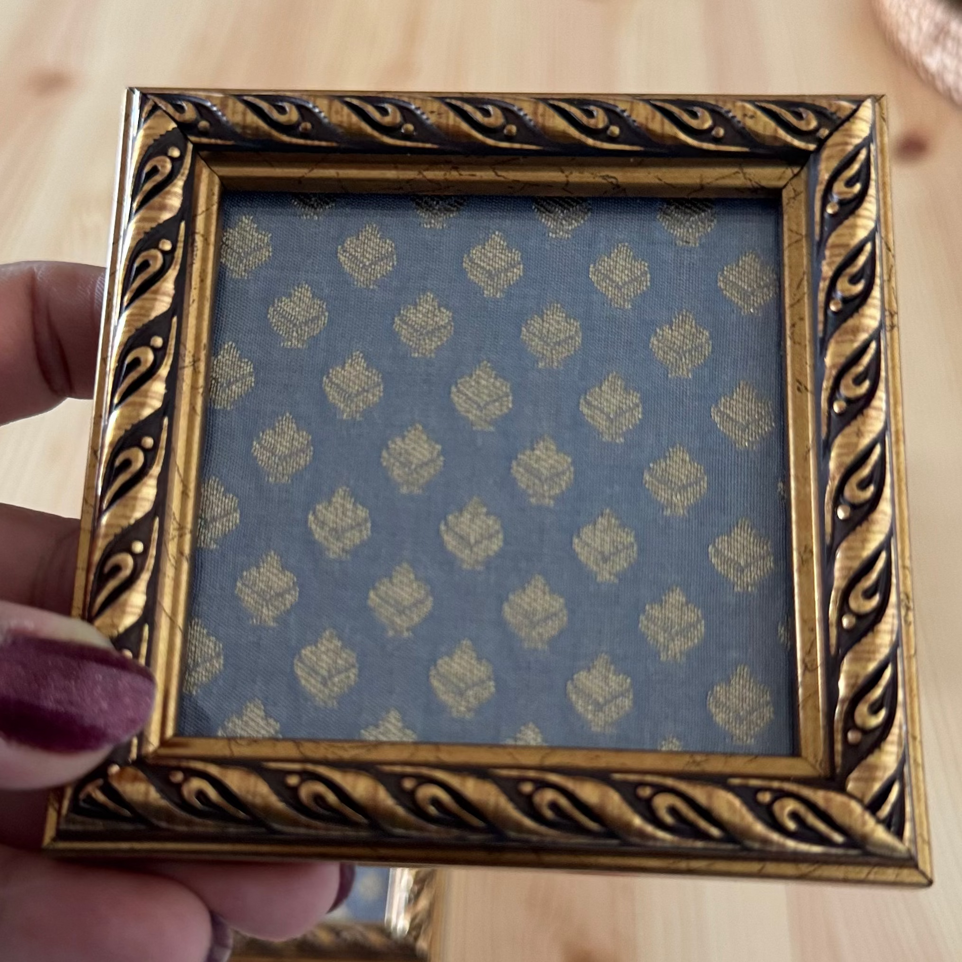 Gold decorative grey brocade fabric coaster held by a hand on a wooden surface.